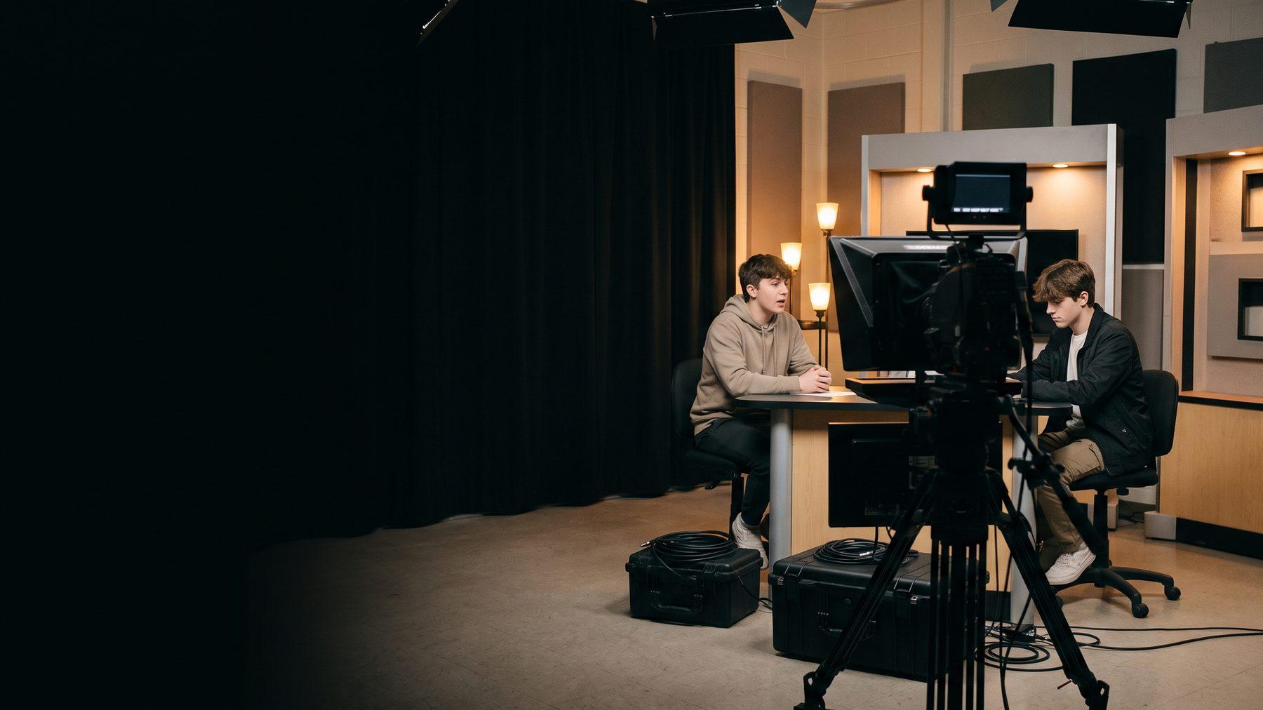 Two students running a school broadcast desk with a camera, teleprompter, and studio lighting